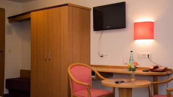 Habitación de hotel con armario de madera, televisor montado en la pared, una mesa con una silla y una mesita de noche con un teléfono y una botella.
