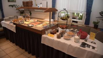 A buffet setup with various food items and utensils, a sign indicating 'Nutella', and a window with a view of greenery.