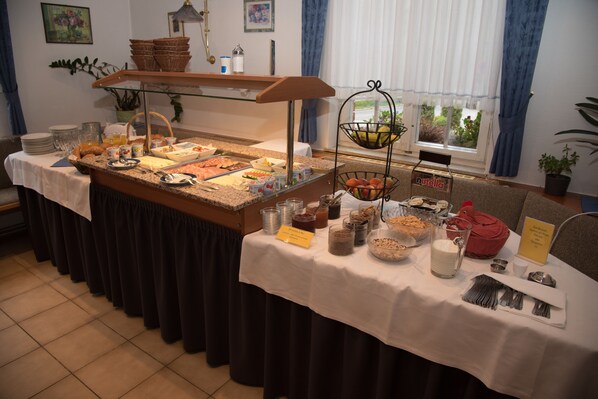 A buffet setup with various food items and utensils, a sign indicating 'Nutella', and a window with a view of greenery.