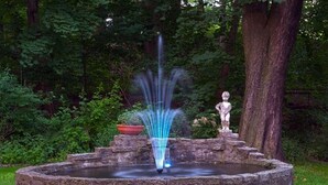 Une fontaine en pierre, dotée d’un jet d’eau central, est entourée d’un bassin circulaire également en pierre, le tout étant installé dans un jardin luxuriant, avec un feuillage dense en arrière-plan.
