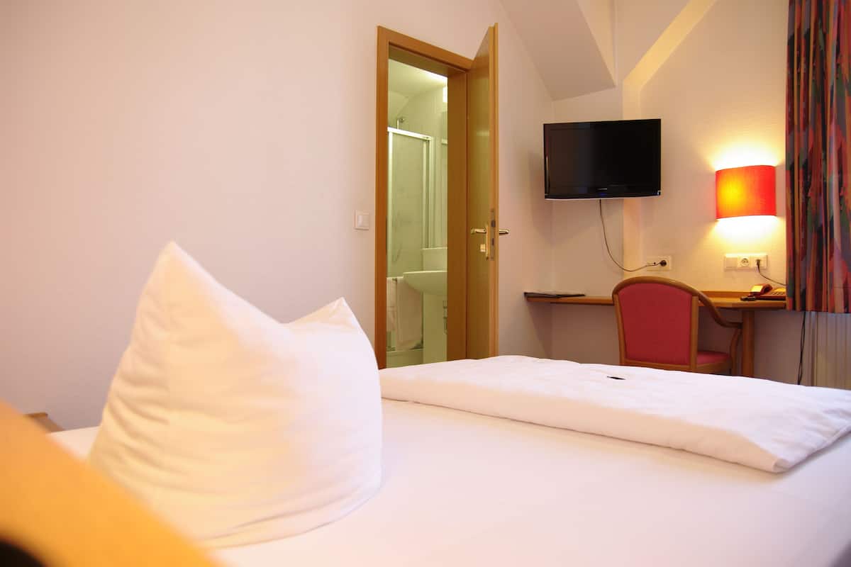 A hotel room with a bed, a desk, a chair, a television, and a shower visible through an open door.