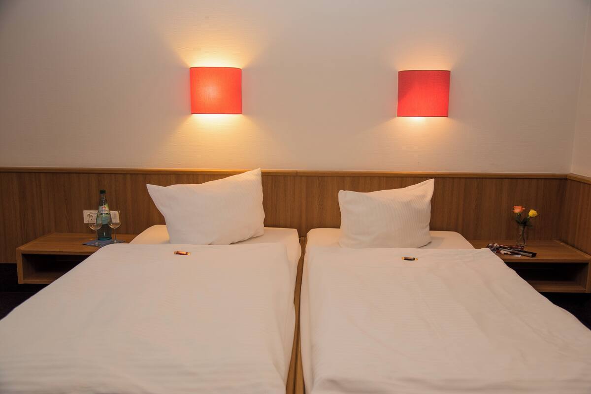 A hotel room with two single beds, wooden headboards, bedside tables with lamps, and a nightstand with a bottle and glasses.