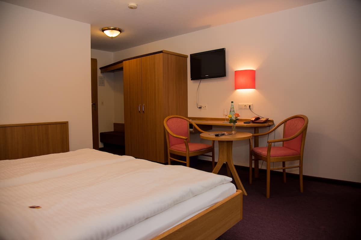 A hotel room with a bed, a desk with two chairs, a television, and a red wall lamp.