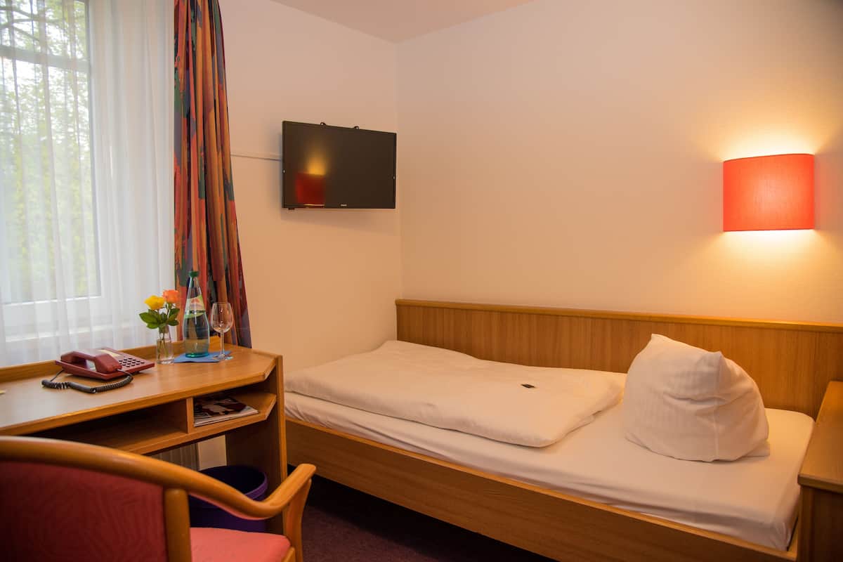 A hotel room with a single bed, a desk with a telephone and a vase with flowers, a television on the wall, and a wall-mounted lamp.