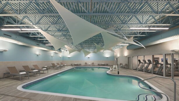 An indoor swimming pool with a modern design, including a high ceiling with a truss structure and overhead lighting. - Courtyard by Marriott Oshawa (Oshawa)