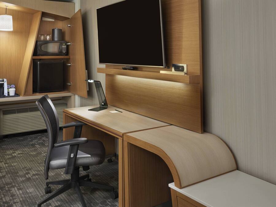 Une chambre d’hôtel compacte comprenant un bureau en bois, un téléviseur à écran plat, un four à micro-ondes, un réfrigérateur, une machine à café et une chaise.