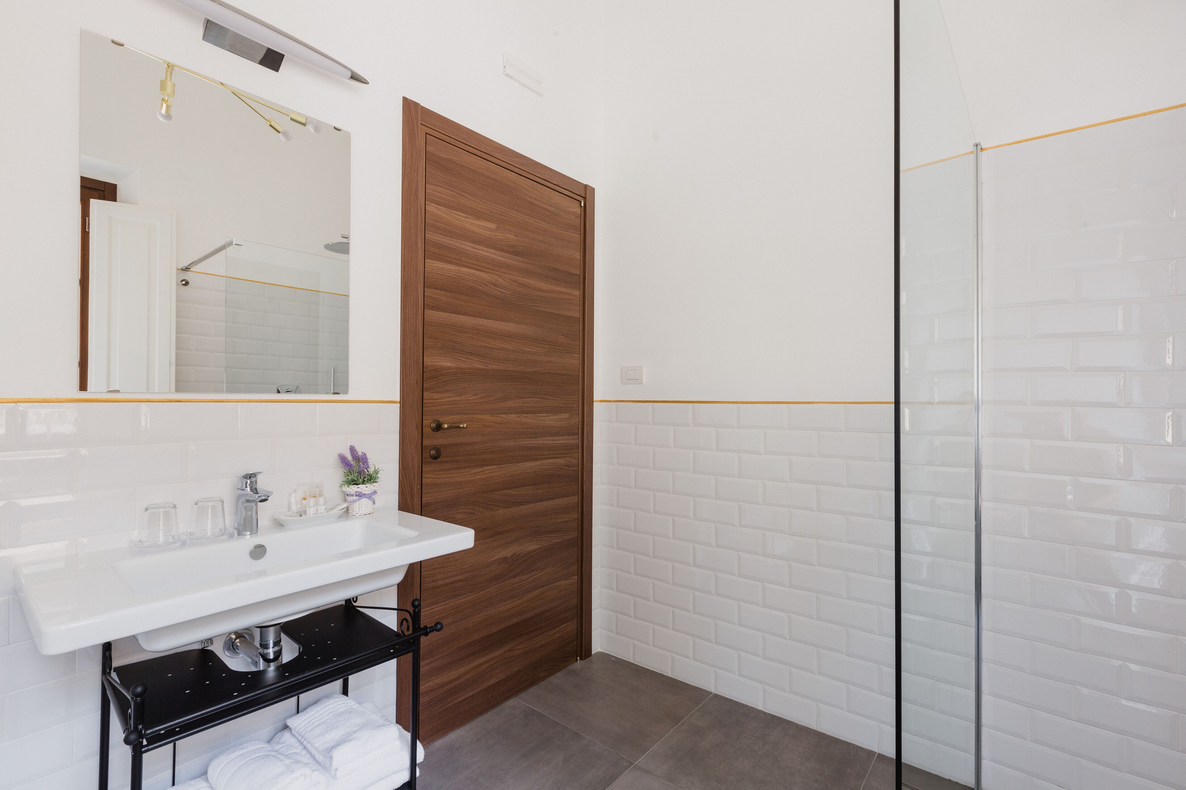 A bathroom with a wooden door, white tiled walls, a white sink, and a glass shower enclosure.