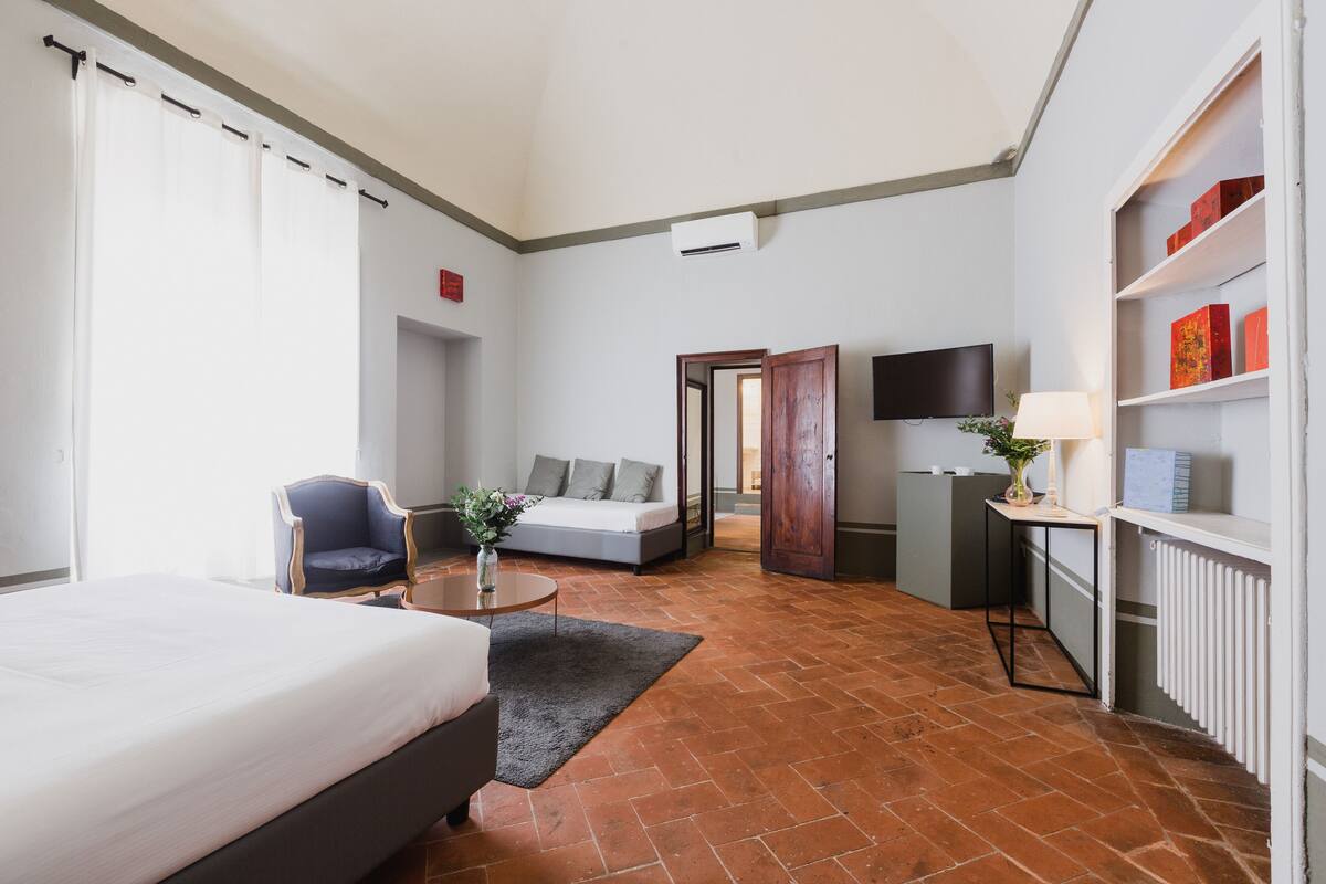 A modern hotel room with a bed, a sofa, a chair, a small table with a lamp, and a TV.