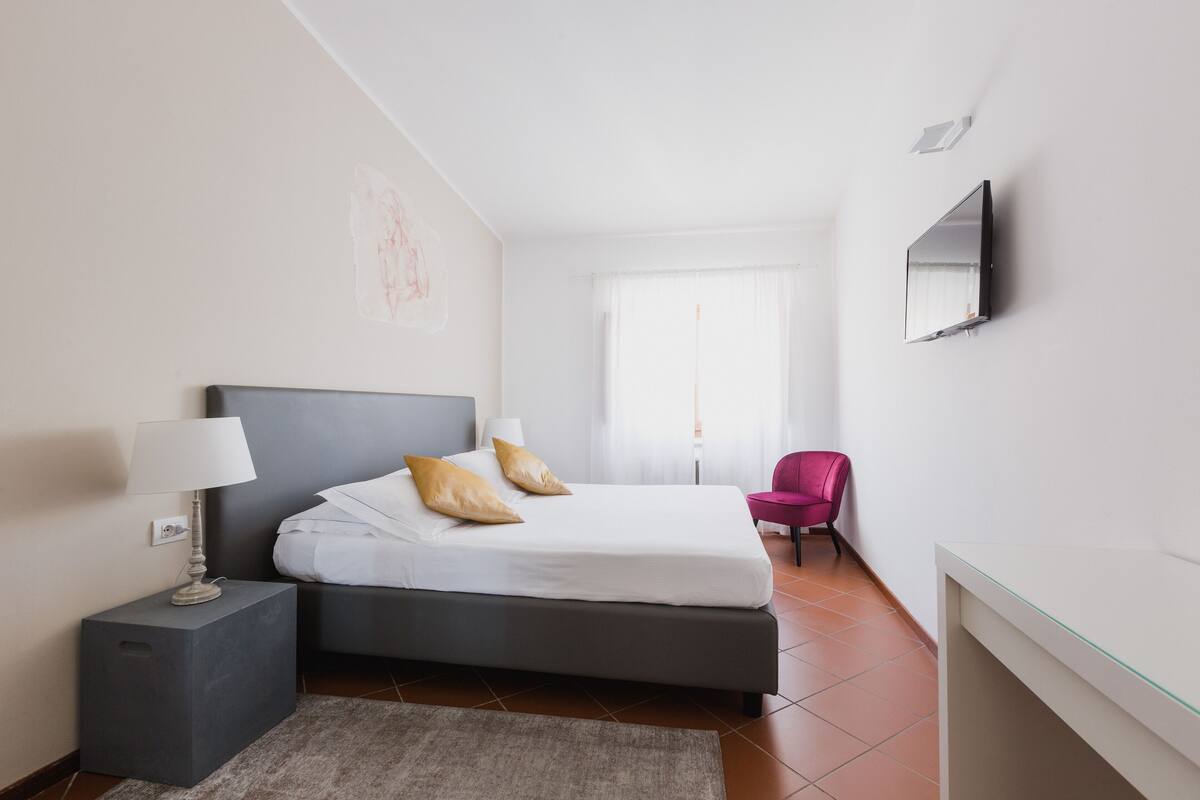 A modern hotel room with a bed, bedside lamp, chair, and a TV mounted on the wall.
