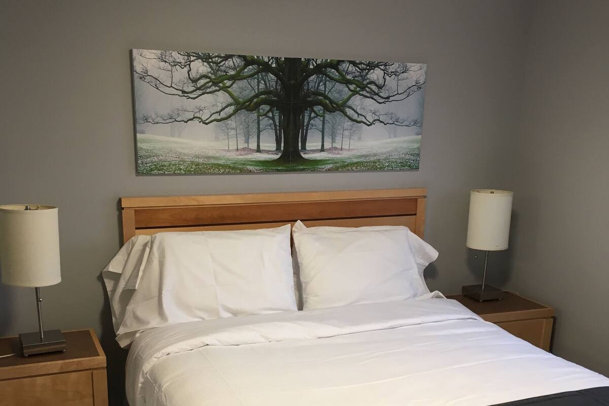 Un lit bien fait, avec du linge de lit blanc, une tête de lit en bois et un plaid noir. Il y a deux tables de chevet avec des lampes, et un tableau représentant un arbre est accroché au mur.