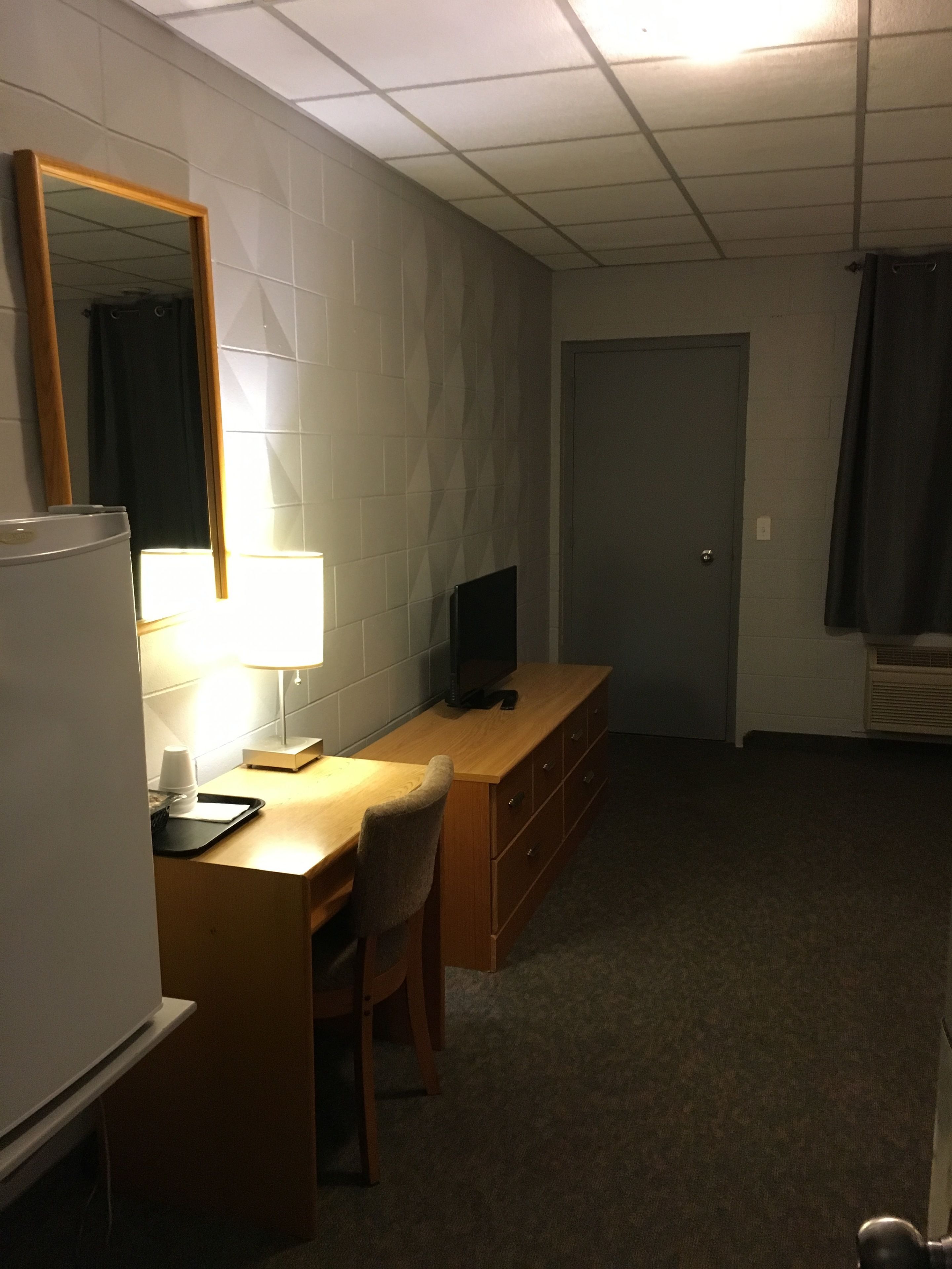 Ein kompaktes Hotelzimmer mit Küchenzeile, einem Holztisch mit Stuhl, einem Fernseher, einem Spiegel und einer Tür.