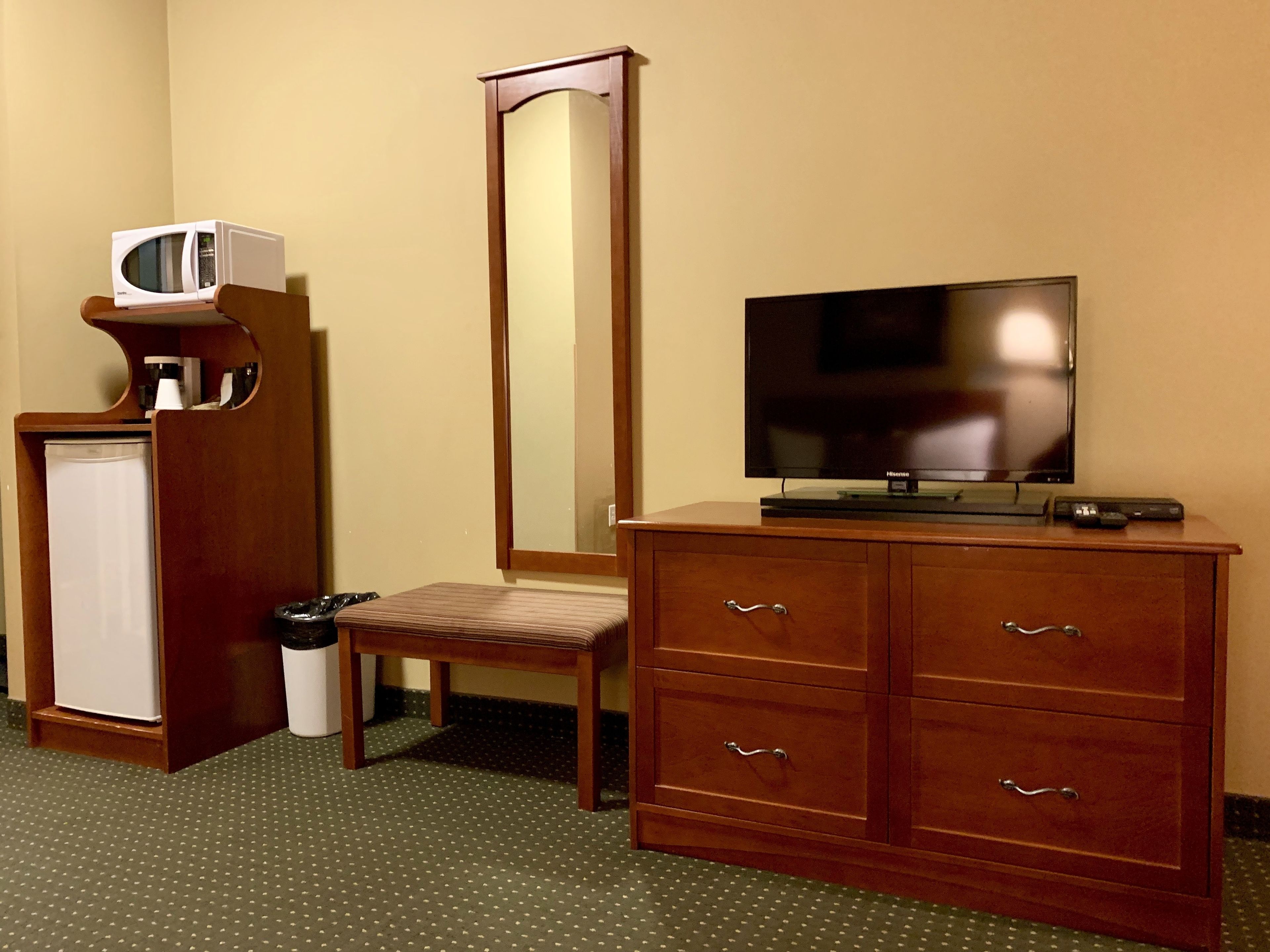 A hotel room with a wooden cabinet, a television, a small refrigerator, a trash bin, and a mirror.