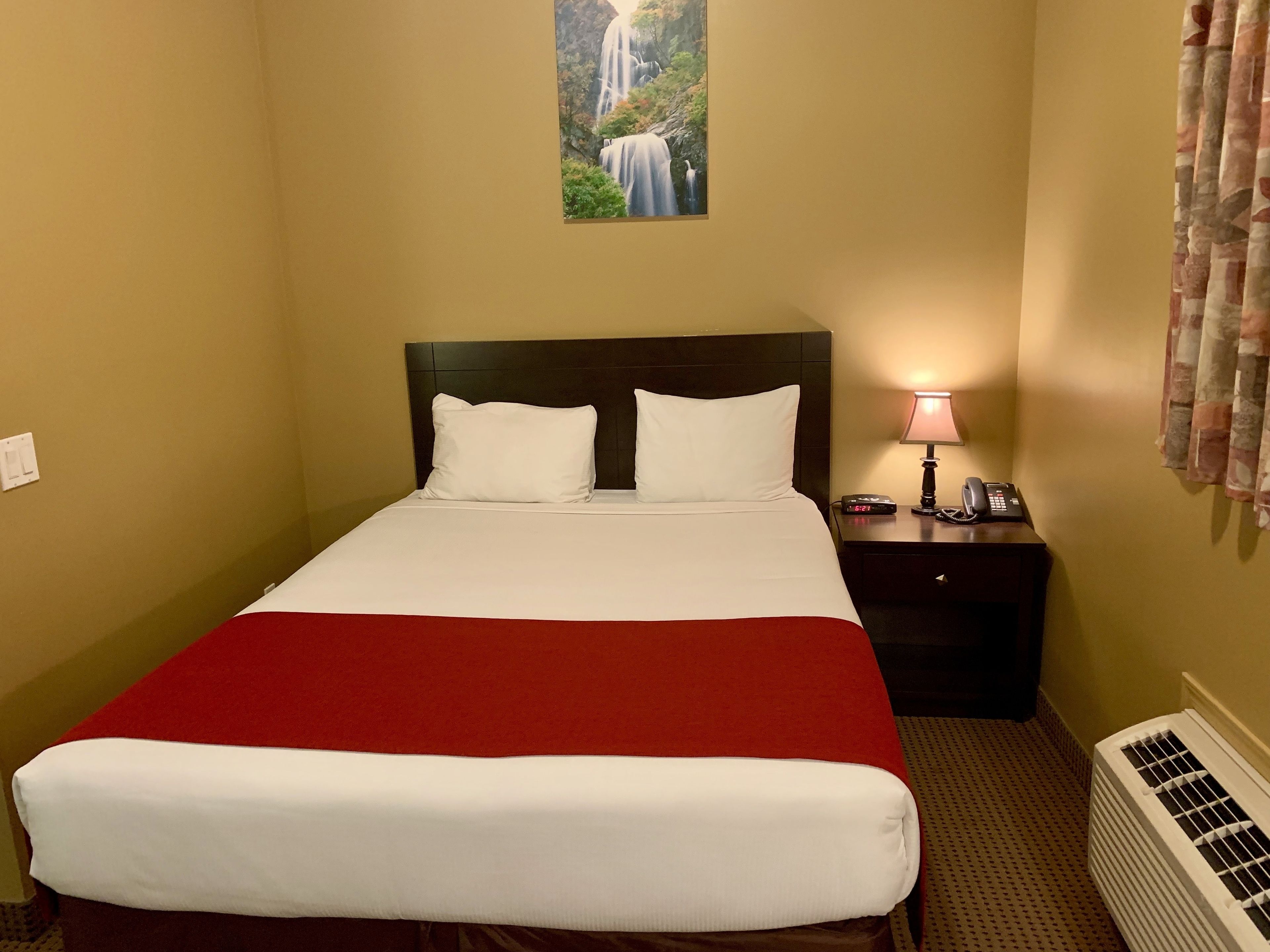 A hotel room with a bed, a nightstand with a lamp and phone, a painting of a waterfall, and a radiator.