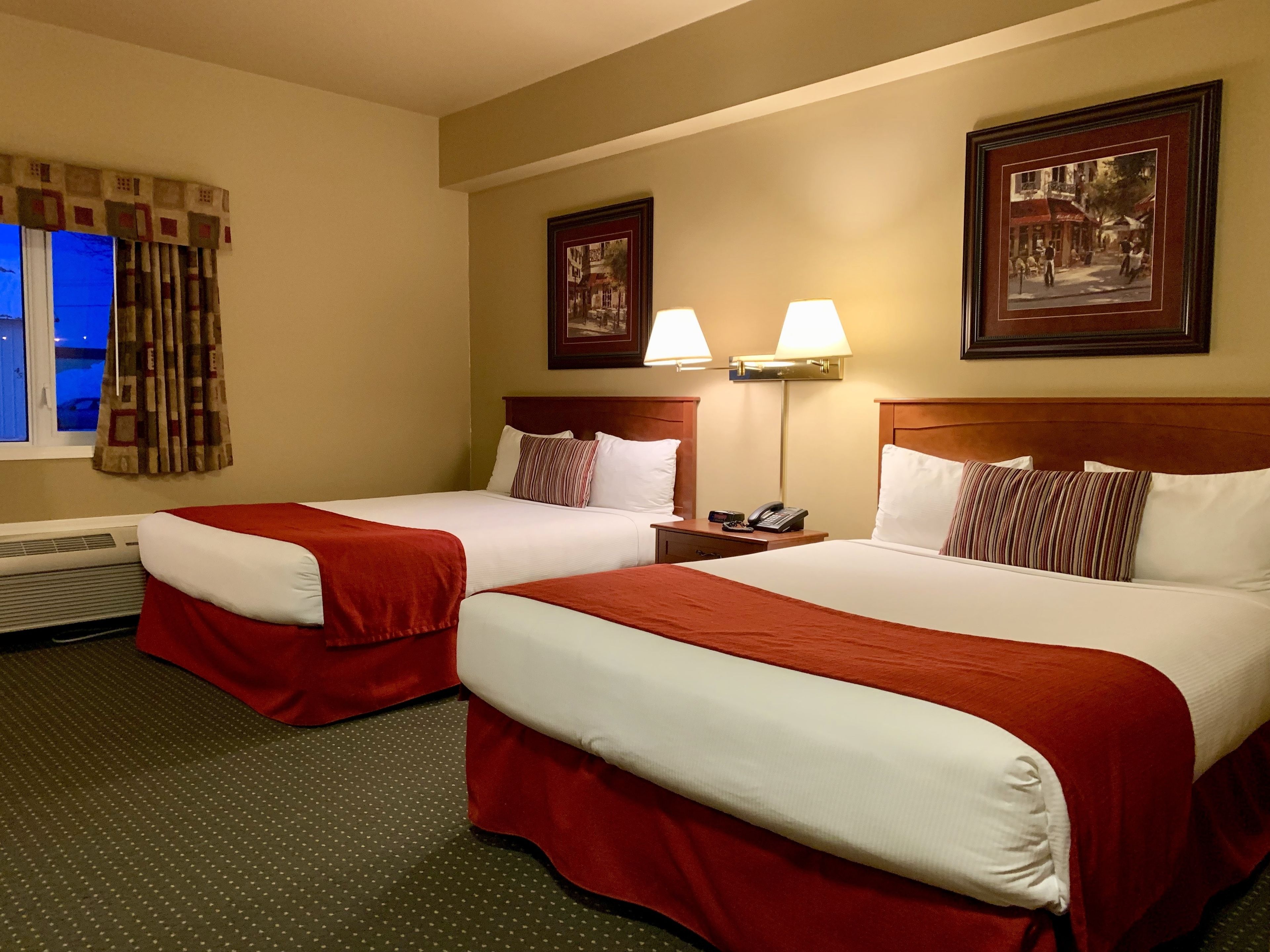 Una habitación de hotel con dos camas, cada una con ropa de cama roja y blanca, una mesita de noche con lámpara y un cuadro en la pared.