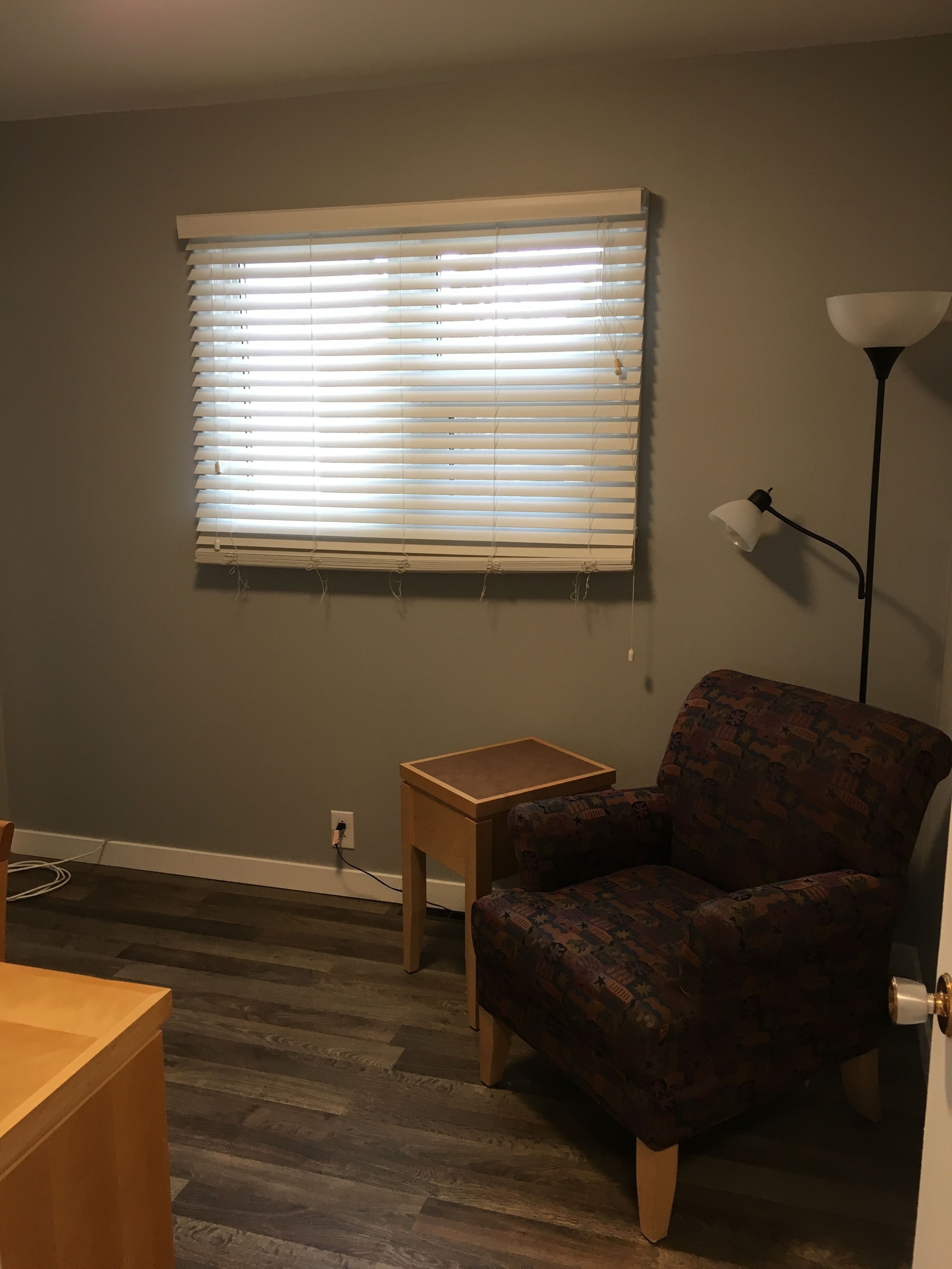 A room with a window with blinds, a patterned armchair, a small wooden table, and a floor lamp.