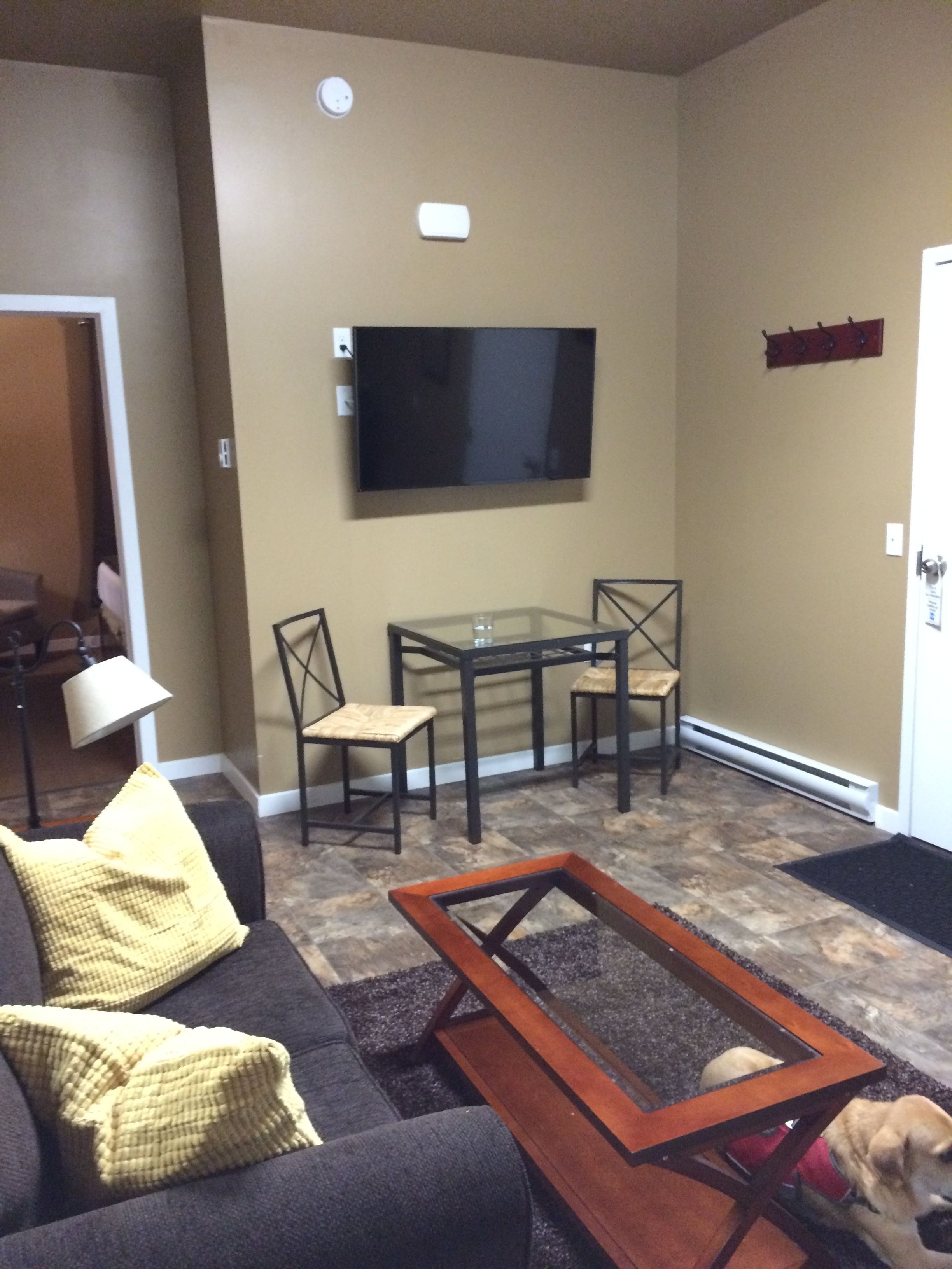 A living room with a dark-colored sofa, yellow cushions, a glass-top coffee table, and a TV mounted on the wall.
