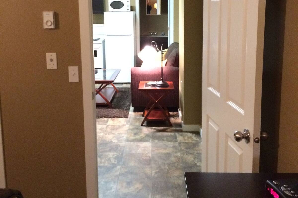 Un couloir mène à un espace de vie comprenant un canapé, une table basse et une lampe. On y trouve un four à micro-ondes posé sur un comptoir, un téléviseur fixé au mur et une porte donnant sur une autre pièce.