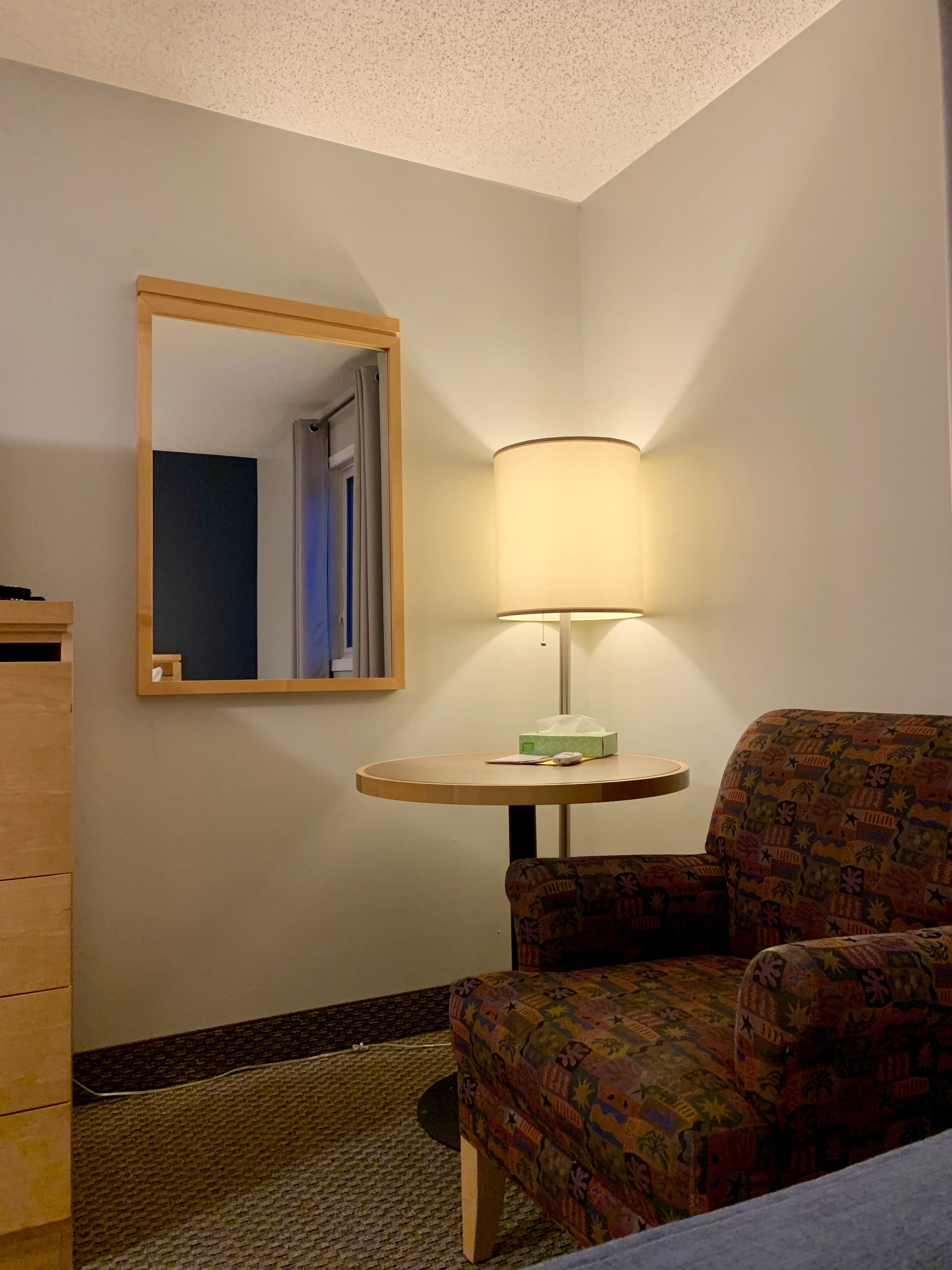 Une chambre d’hôtel avec un lit simple, une chaise, une petite table avec une lampe, un miroir et une commode.