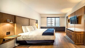 Quarto de hotel moderno com uma cama grande, uma televisão fixada na parede e uma janela com vista para a cidade.