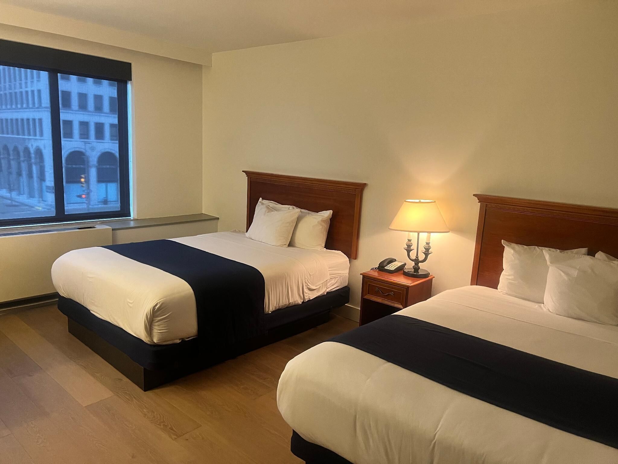 Habitación de hotel con dos camas, una mesita de noche con lámpara y ventana con vistas a edificios.