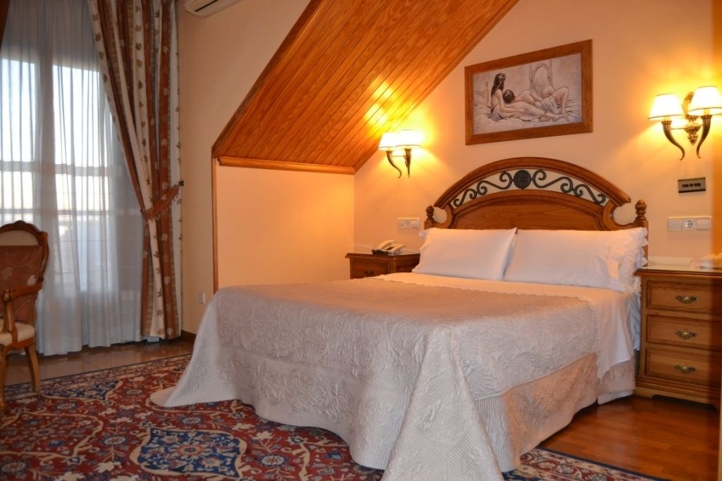 Une chambre avec un plafond en bois, un grand lit recouvert de draps blancs, deux lampes fixées au mur et un tapis à motifs.
