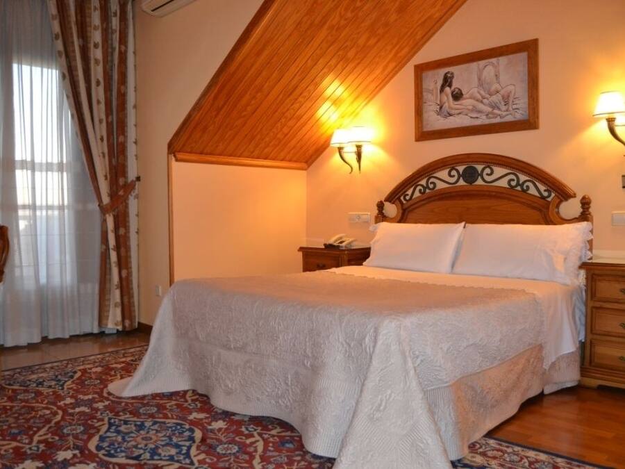 Une chambre avec un plafond en bois, un grand lit recouvert de draps blancs, deux lampes fixées au mur et un tapis à motifs.
