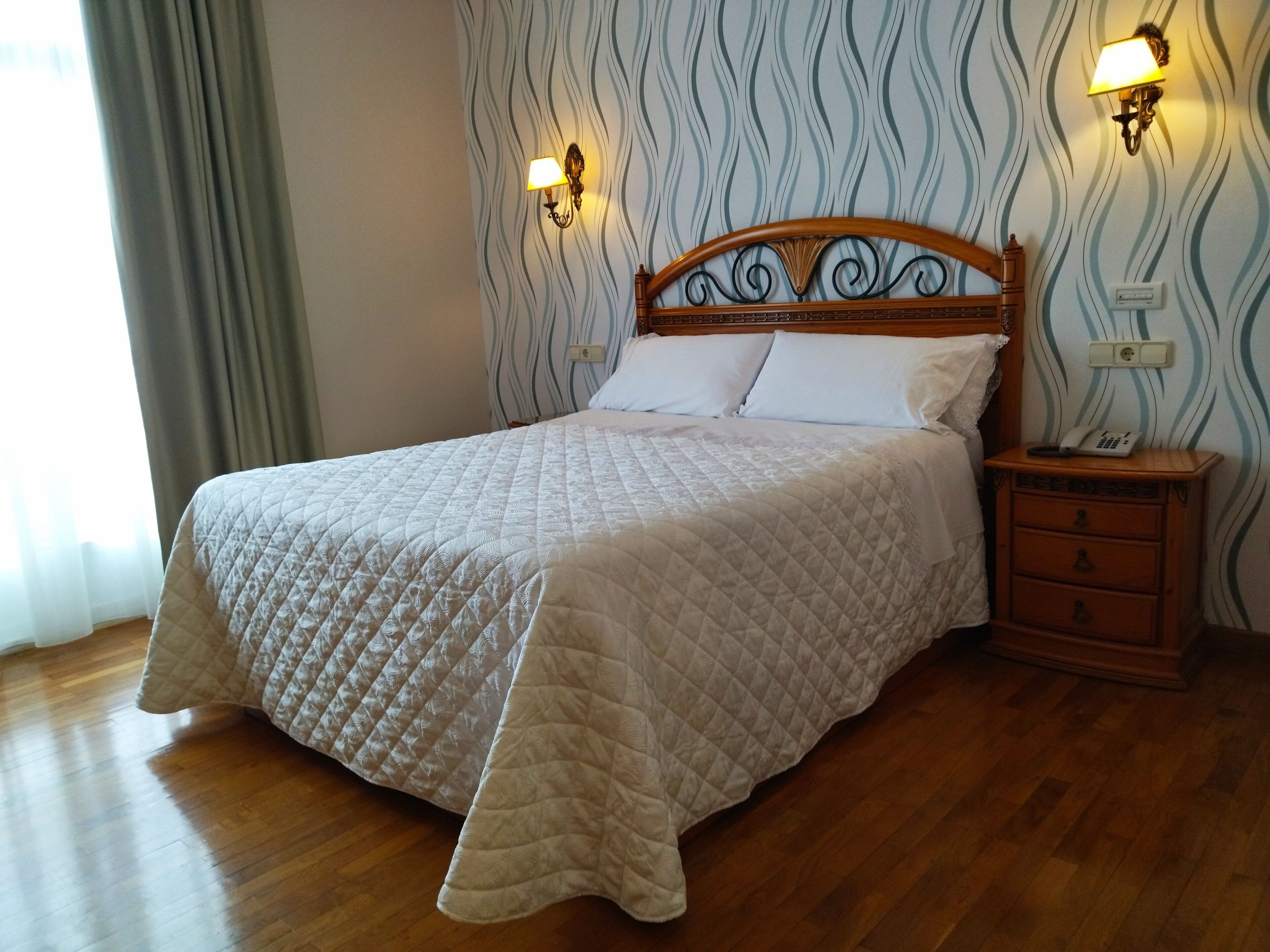 A neatly made bed with a quilted cover, a wooden headboard, and two wall-mounted lamps.