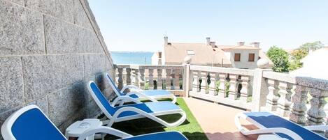 A terrace with blue lounge chairs and a stone wall, overlooking a coastal area with buildings.