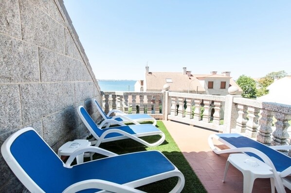 A terrace with blue lounge chairs and a stone wall, overlooking a coastal area with buildings. - Hotel Playa Compostela (Vilagarcia de Arousa)