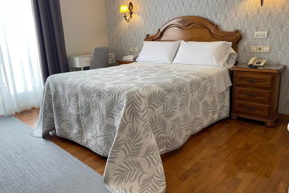 Ein ordentlich bezogenes Bett mit gemustertem Überwurf, einem Holz-Kopfteil, Nachttischlampen, einem Nachttisch und einem Telefon, sowie Tapete mit Muster.