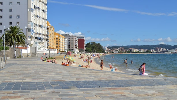 Una playa con gente tomando el sol y nadando, un sendero de piedra y edificios al fondo.