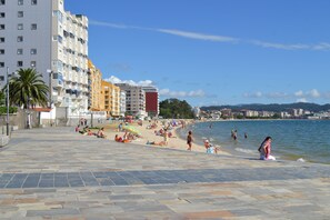 Een strand met mensen die zonnebaden en zwemmen, een pad van stenen en gebouwen op de achtergrond.