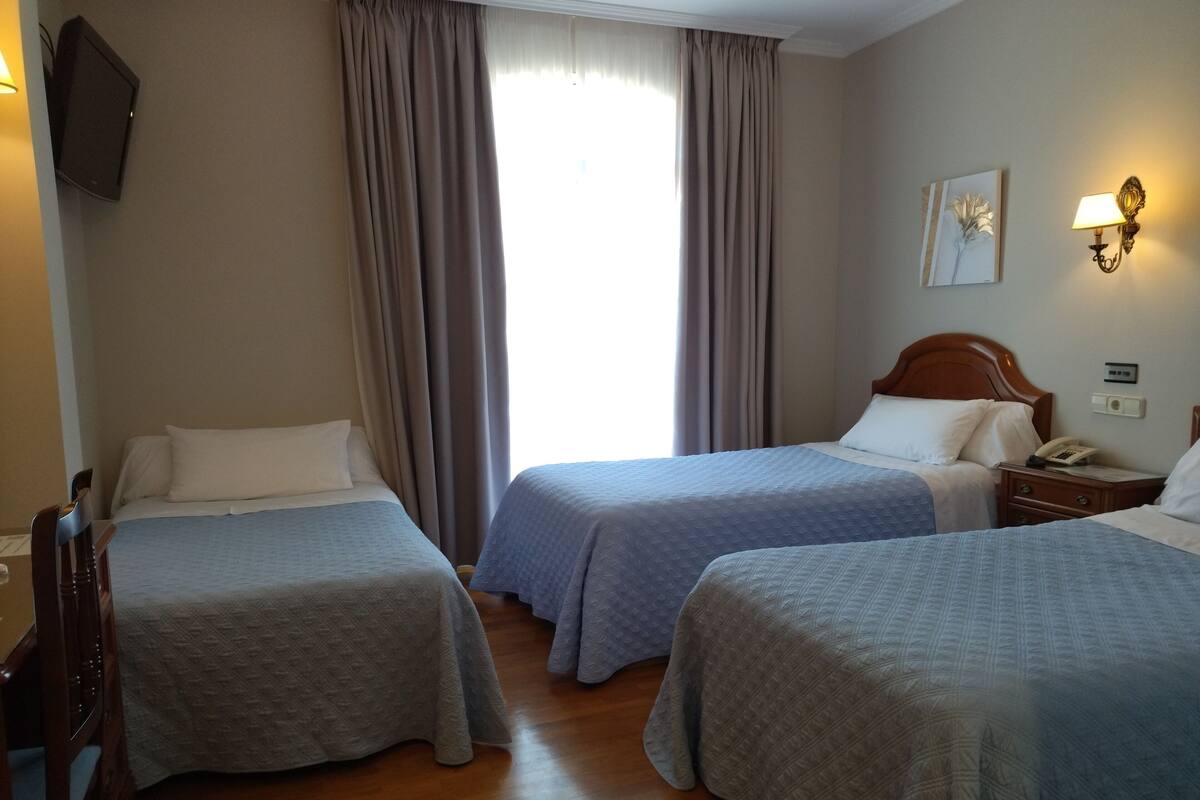 Ein Hotelzimmer mit zwei Betten, einem Holzsessel, einem Nachttisch mit Lampe und einem Fenster mit Vorhängen.