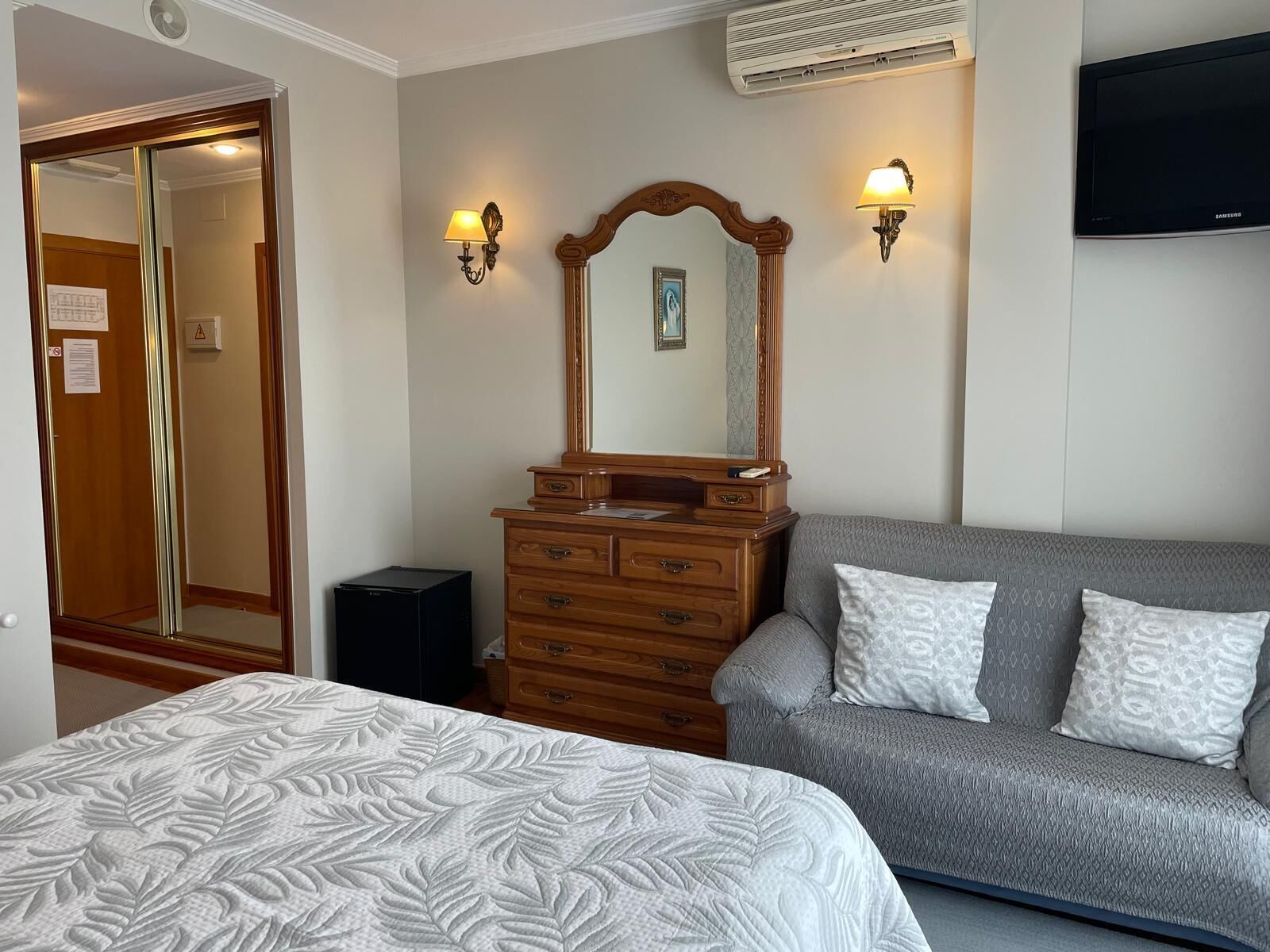 A bedroom with a bed, a dresser, a gray sofa, two wall-mounted lamps, and a flat-screen TV.