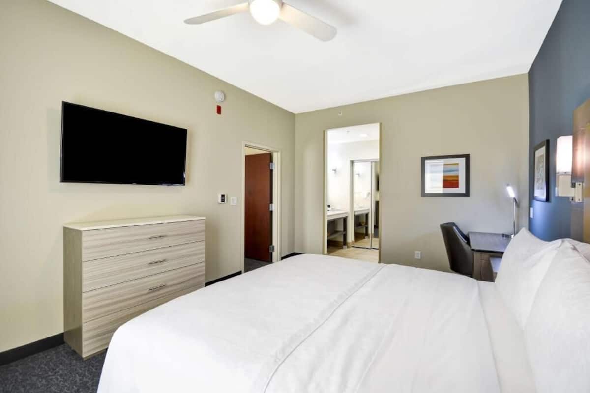 Una habitación de hotel con una cama, televisión montada en la pared, escritorio y una silla.