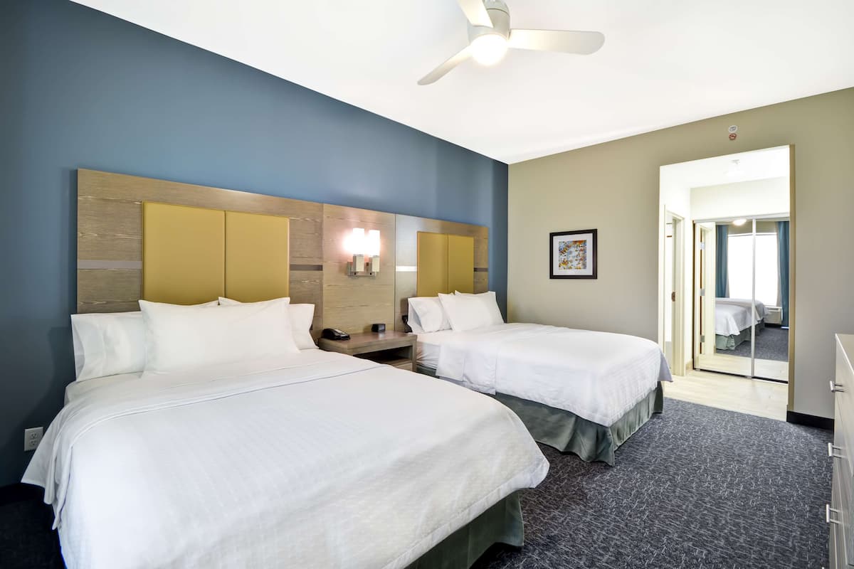Habitación de hotel con dos camas, un ventilador de techo, una mesita de noche y una puerta que da a otra habitación.