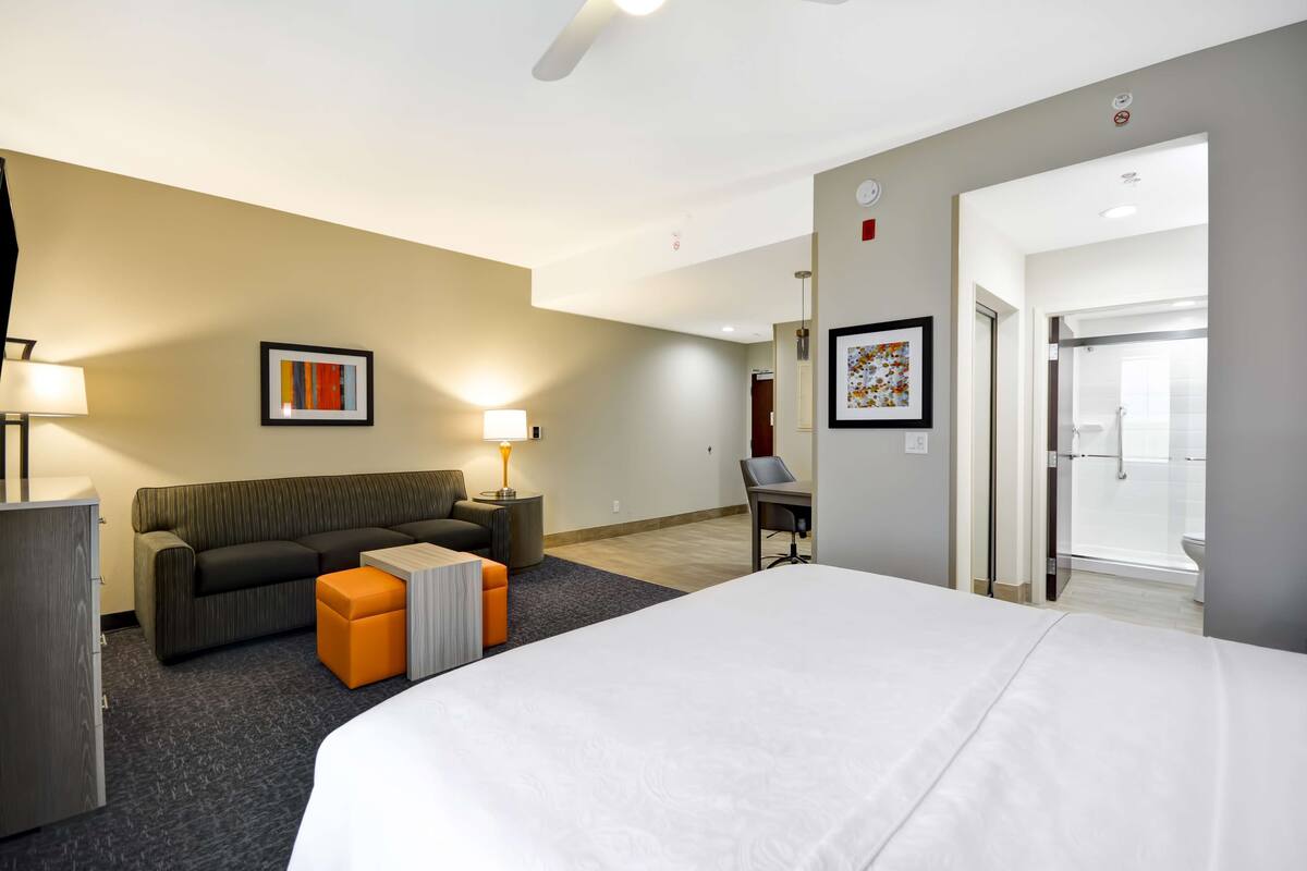 Habitación de hotel con una cama grande, un sofá, una mesita, una lámpara y un escritorio. Se ve un baño por una puerta abierta.