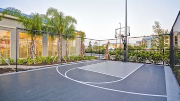 Un terrain de basket en plein air, entouré d’un filet de protection, bordé de palmiers et de bâtiments résidentiels.