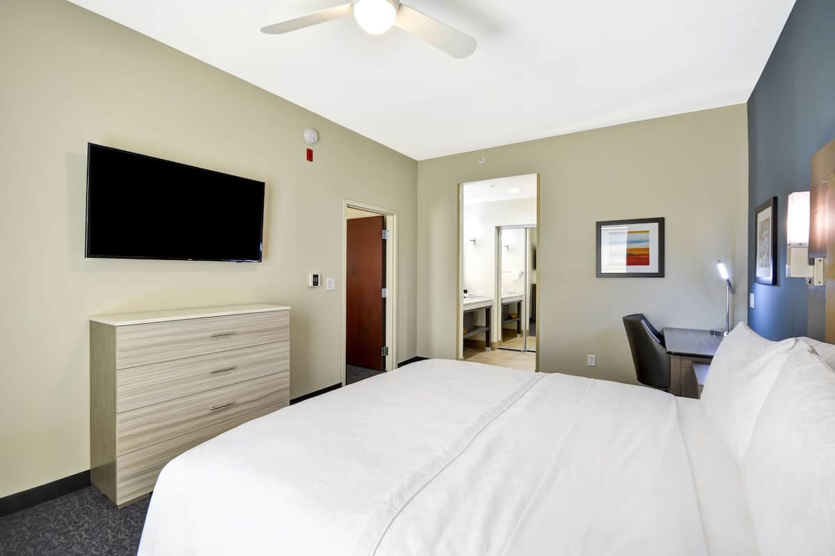 Habitación de hotel con cama, escritorio, silla, televisión y baño visible a través de una puerta abierta.