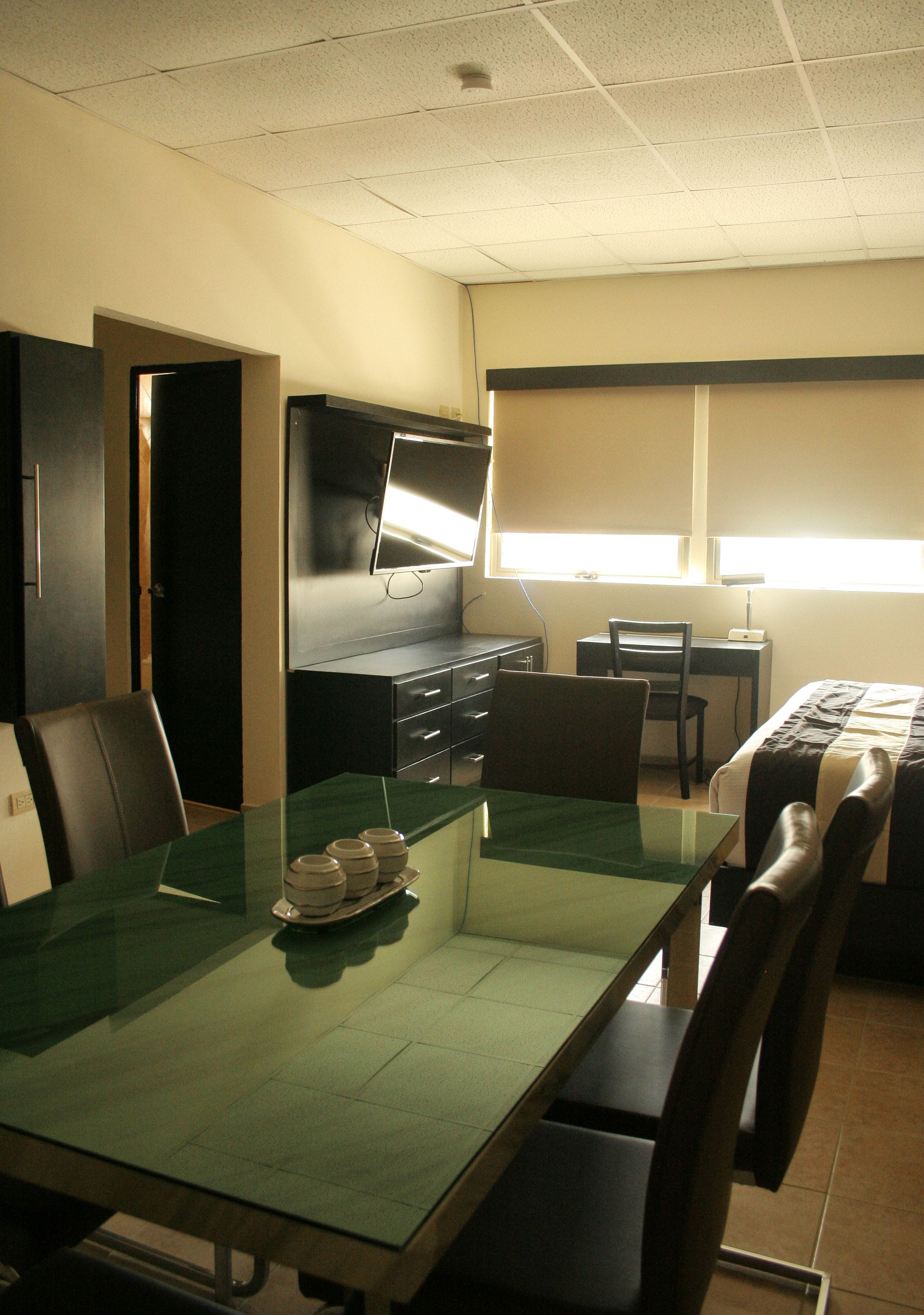 A hotel room with a glass dining table, a TV mounted on the wall, a desk with a chair, and a bed with a patterned bedspread.