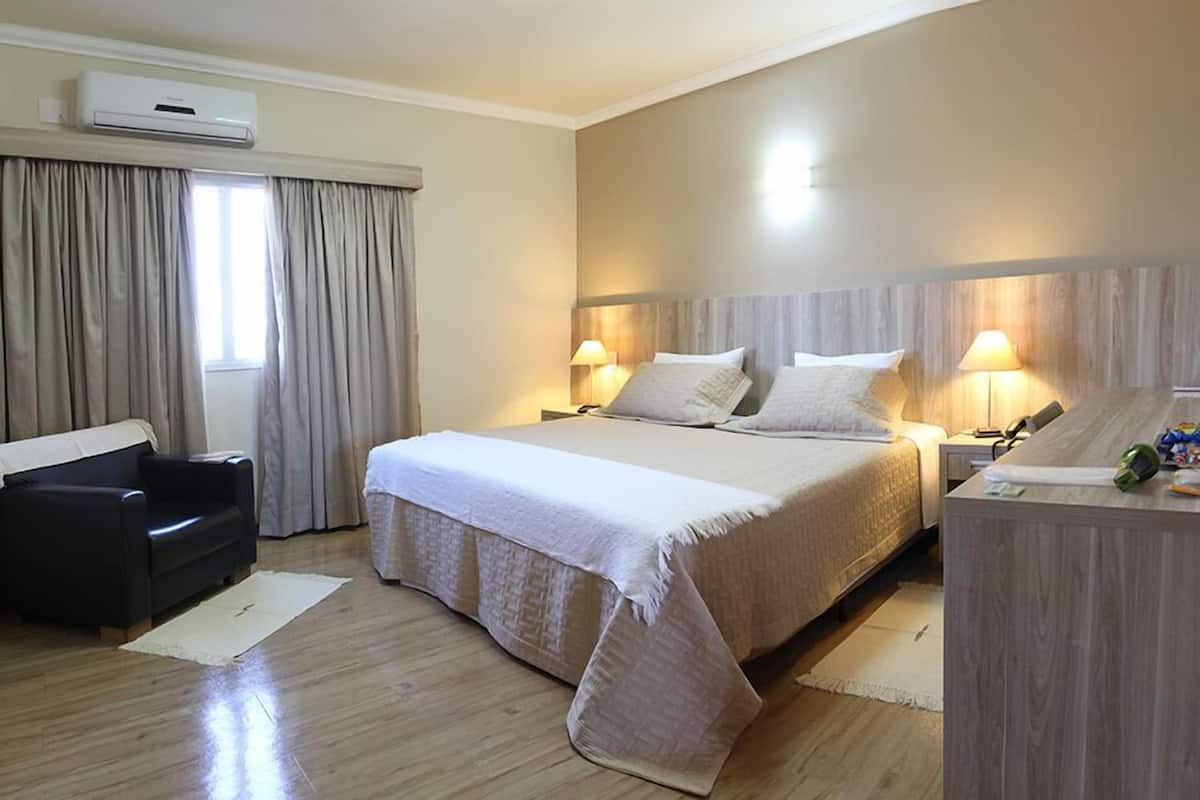 A hotel room with a large bed, two bedside tables with lamps, a desk, a chair, and an air conditioning unit.