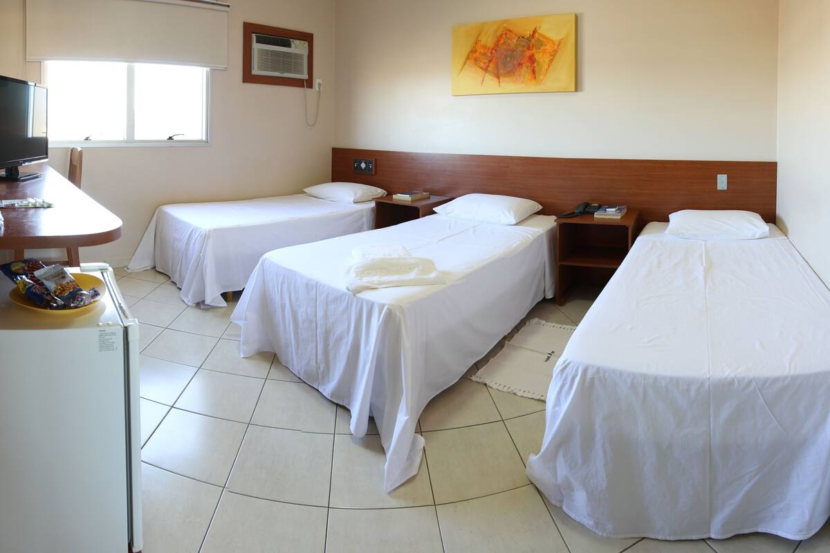 A hotel room with two single beds, a desk with a computer, a small table with a bowl of fruit, and a window providing natural light.