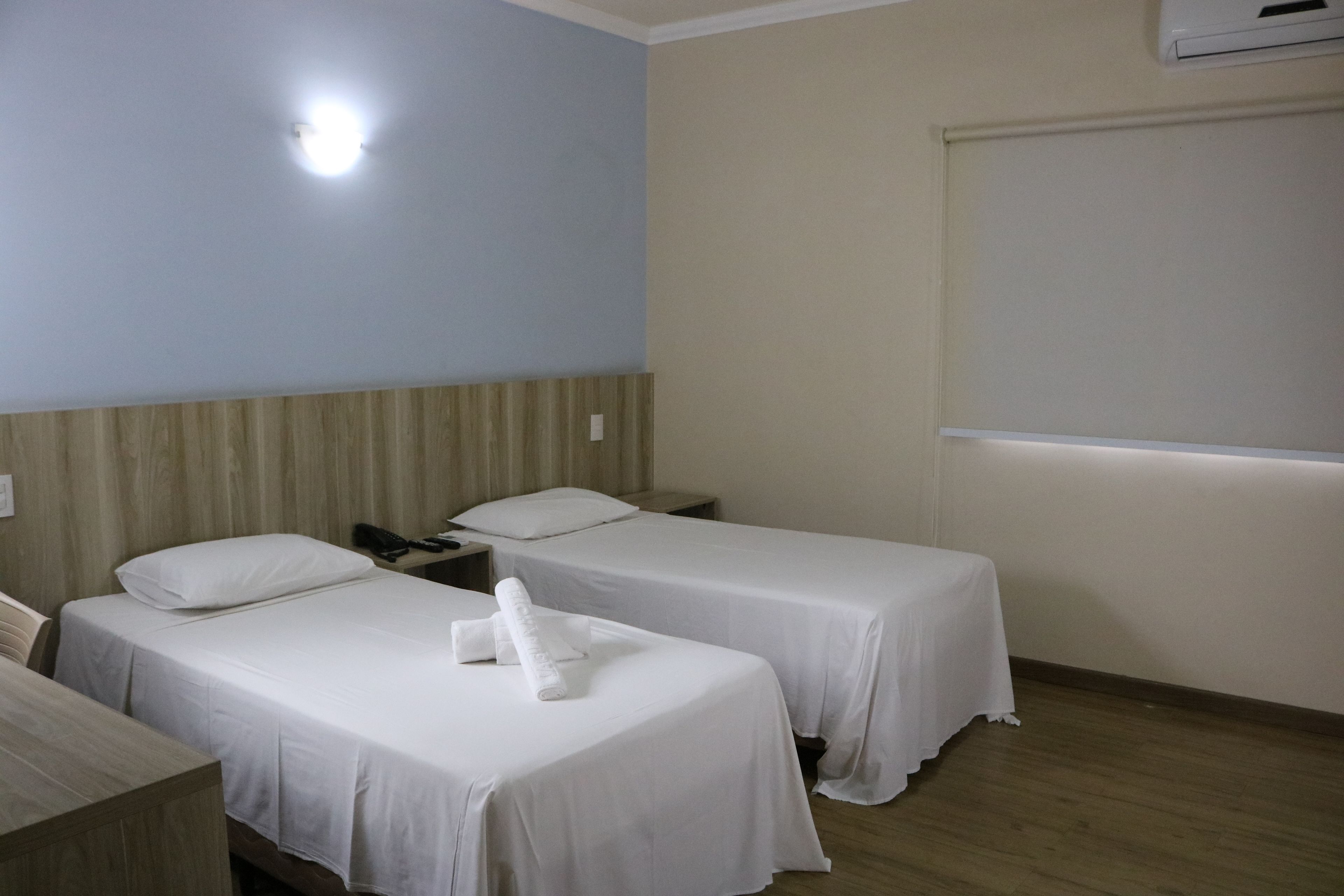 A hotel room with two single beds, a wooden headboard, a bedside table with a lamp, and a window with a blind.