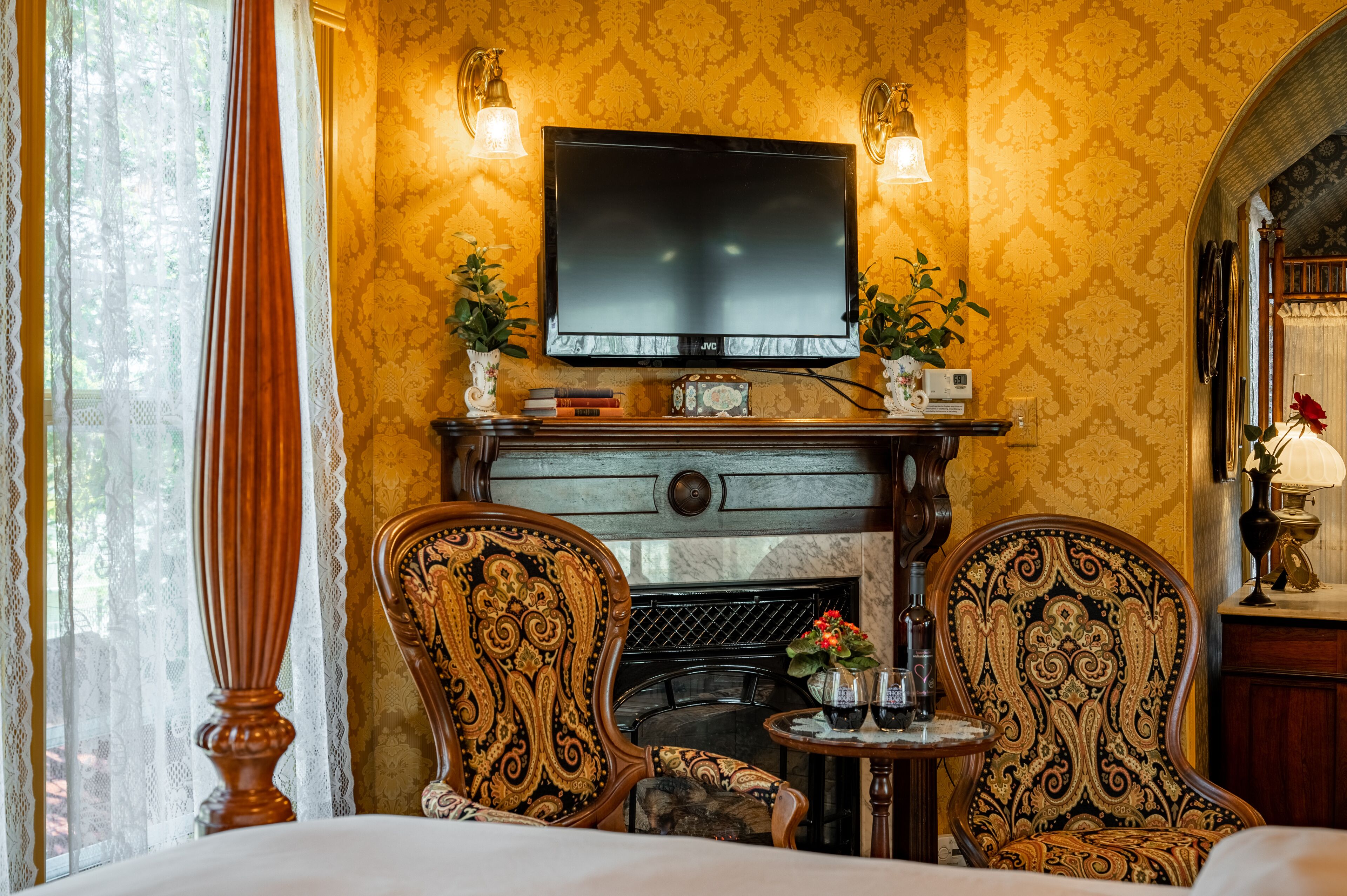 A room with a television mounted above a fireplace, two ornate chairs, a small table with a vase, and a window with curtains.
