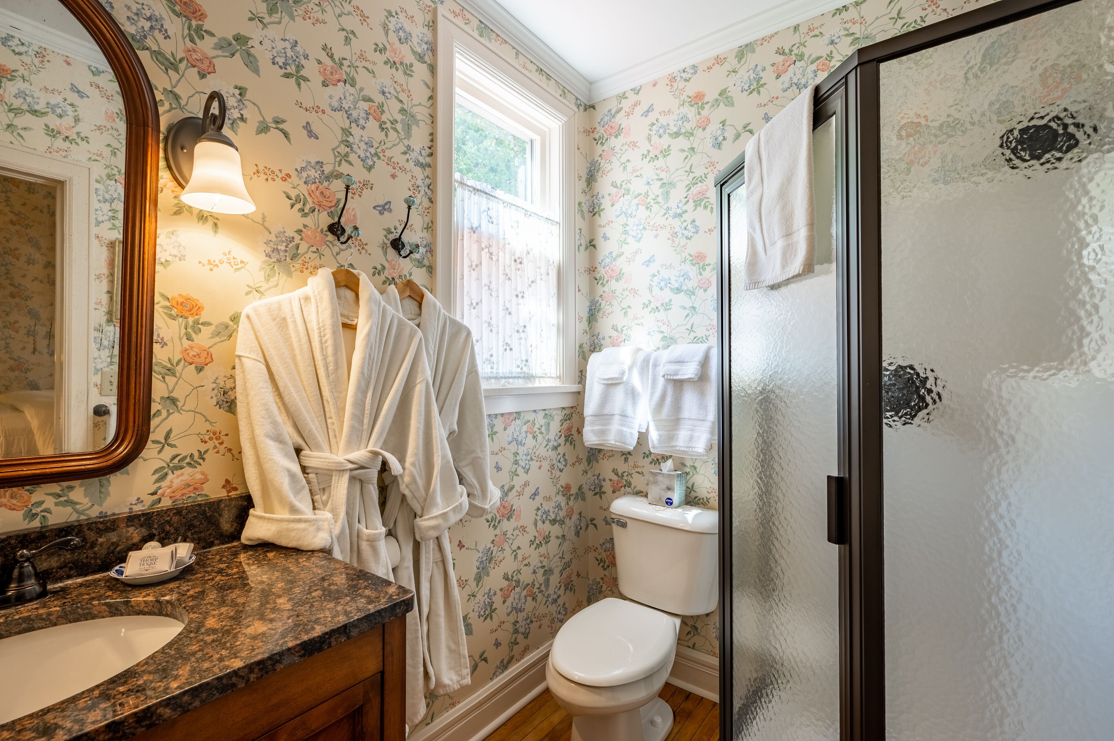 A bathroom with a floral wallpaper, a large mirror, a toilet, and a shower area.