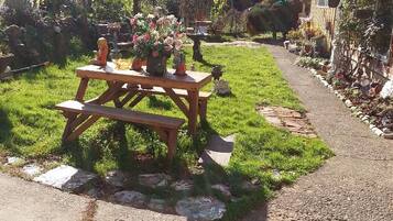Un jardín con una mesa de picnic de madera y bancos, rodeado de diversas plantas y árboles.