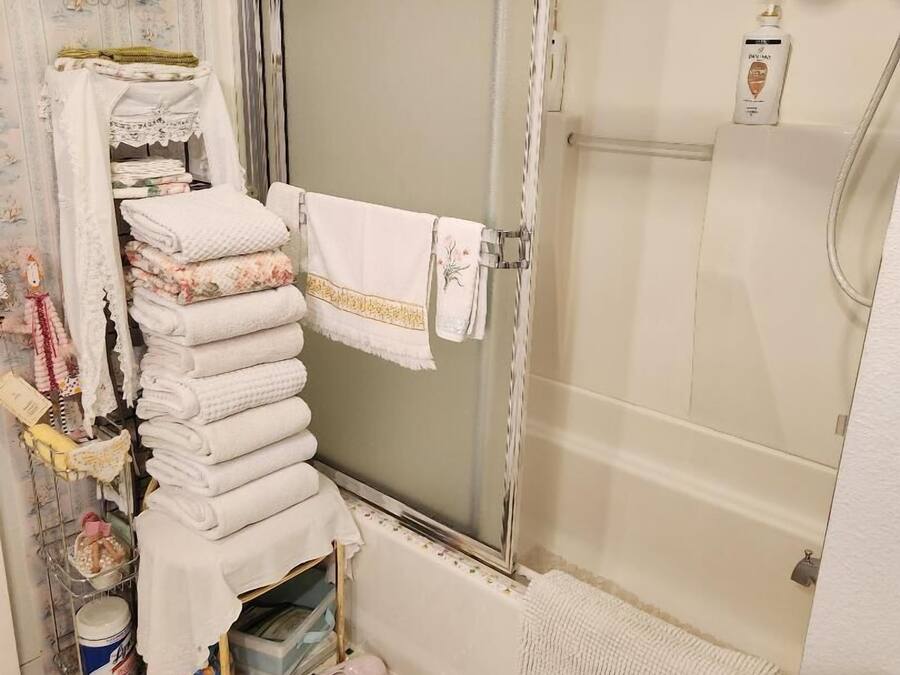 A bathroom with a shower, a towel rack with folded towels, a decorative wall hanging, and a small shelf with toiletries.