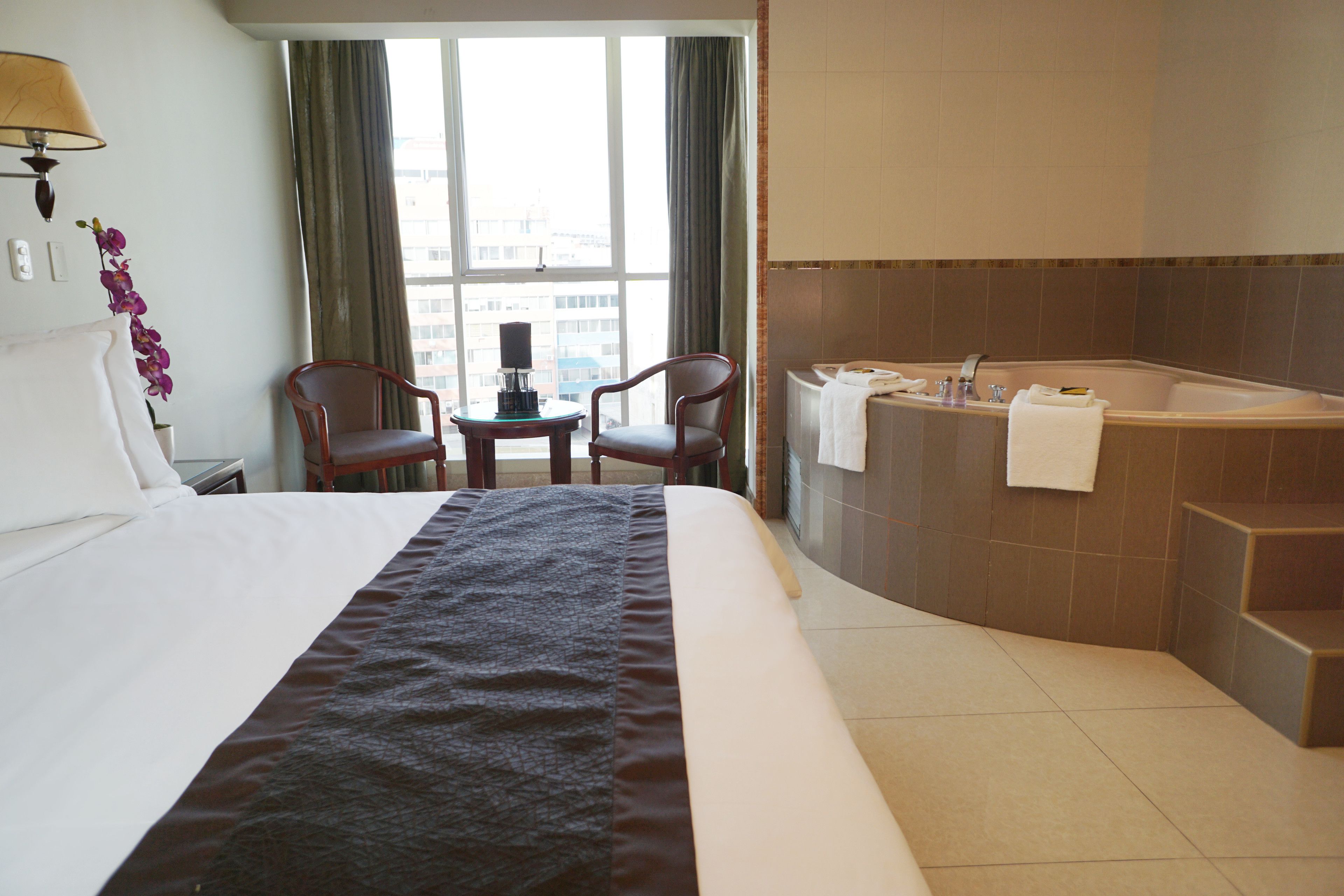 A hotel room with a large bed, a sitting area with two chairs, a small table with a lamp, a window with curtains, and a bathroom with a bathtub and towel rack.