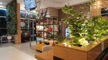 Un moderno vestíbulo de hotel con un mostrador de recepción, una planta grande, un televisor montado en la pared y una estantería con varios objetos.