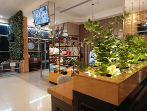 En modern hotellobby med en receptionsdisk, en stor växt, en väggmonterad TV och en hylla med diverse föremål.
